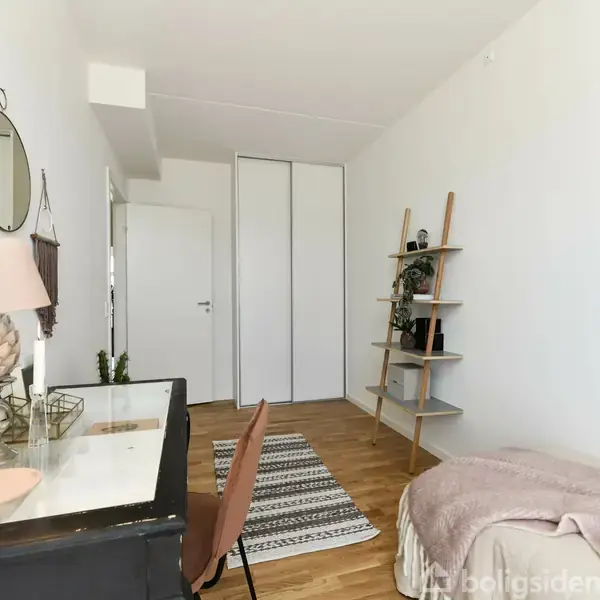 Et skrivebord med lampe og stol står ved en hvid væg. Værelsesskydedøre og en reol med planter i baggrunden. En seng med tæppe står ved siden. Gulvet er trægulv.