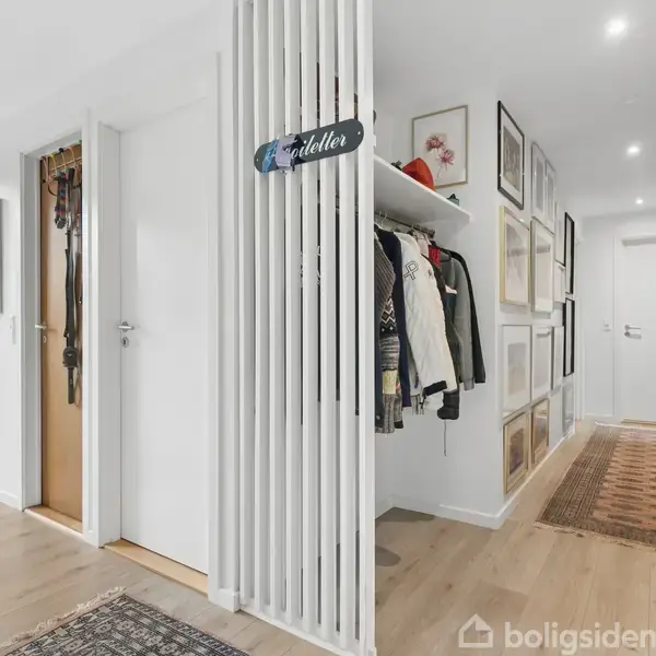 En lys gang med trægulv har frakker hængende på en åben garderobe til højre. Væggene er pyntet med billeder. En dør fører ud til en have med grønne buske. Skilt med teksten "Toilettet".