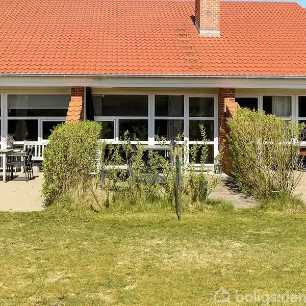 Rækkehus med rød tag og store vinduer. Terrasse med havemøbler og parasol. Omkranset af grønne buske i en velplejet have.
