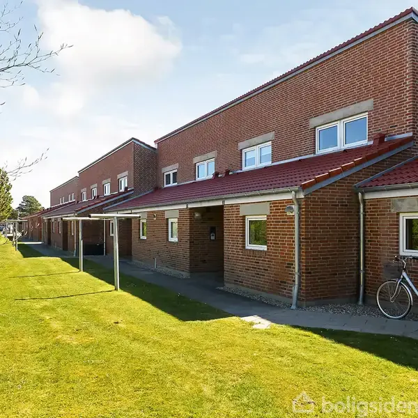 Rødt rækkehus med cykel parkeret foran. Husene har røde tegltage og ligger ved siden af en grøn græsplæne med træer langs venstre side.