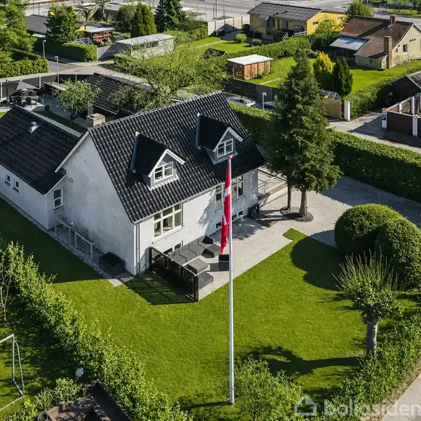 Et hvidt hus med sort tag står på en velplejet græsplæne. Foran huset er der et flag på en høj flagstang. Omkring er der træer og buske i en beboelsesområde.