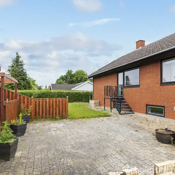 Rød murstensbygning med trappe fører til have, omgivet af hegn og potteplanter, på grå belægningssten. Baggrund med grønne buske og bygninger under delvis overskyet himmel.
