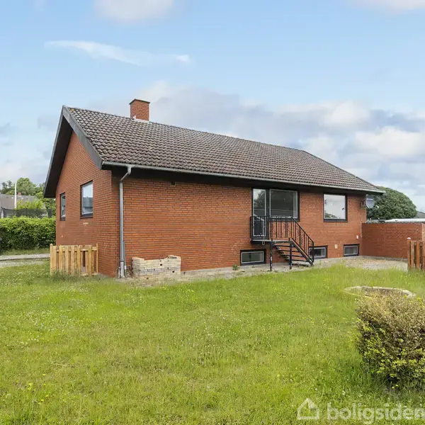 Et rødt murstenshus står i en græsplæne. Der er indgangstrin med rækværk foran huset. I baggrunden er der hække og et klart blåt himmel.