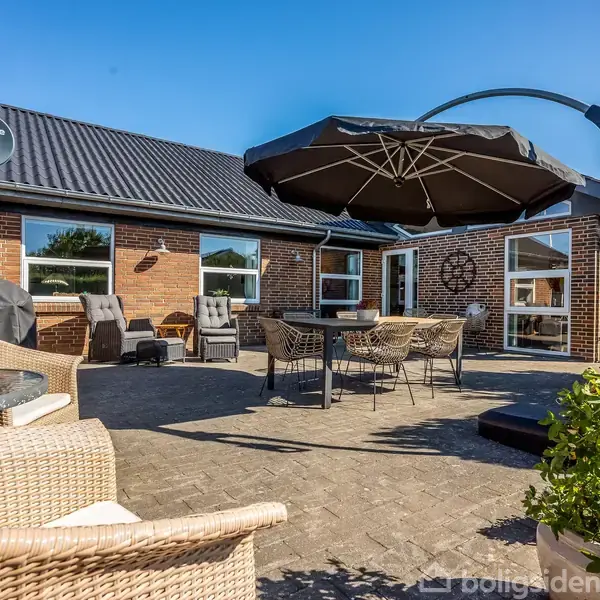 Terrasse med wicker-møbler og stor parasol. Omkranset af murstenshus med sorte tagsten. Himmel er klar og blå. Grønne planter tilføjer farve til miljøet.