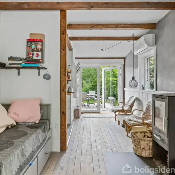 En hyggelig stue med træbjælker, hvor en sofa fyldt med puder står langs væggen. Et brændeovn står til højre, mens døre fører ud til en have med møbler.