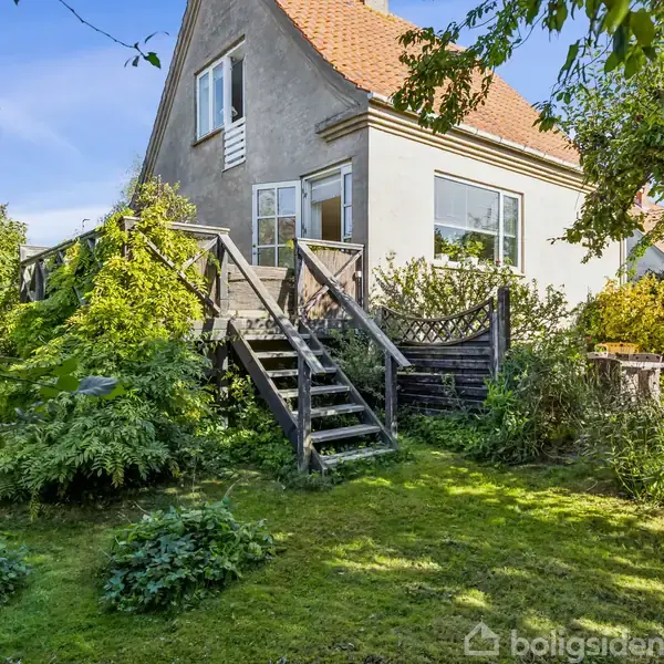 Hus står stille i solskinsfyldt haveomgivelser. Trappe fører op til døren. Omkranset af frodig vegetation og blå himmel.