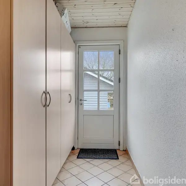 En lys gang leder op til en glasdør med udsigt til en have. Skabsdøre er til venstre, væg til højre. Flisegulvet er lyst, og loftet er træbeklædt.