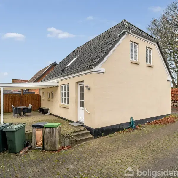 Et beige hus med skråt sort tag står i en gårdhave. I gårdhaven er der affaldscontainere og et halvtag med havemøbler. Omkring ejendommen ses en brostensbelagt gårdsplads.