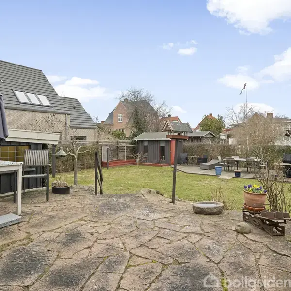 Terrasse med borde og stole står på et stenbelagt område. I baggrunden ses en have med græs, buske og et udhus. Nabohuse omgiver stedet under en blå himmel.