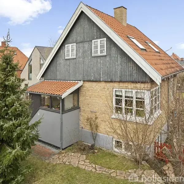 Et hus med rødt tegltag og gul murstensfacade står i en have med græs og buske. Et grantræ vokser tæt ved huset. Himmel med skyer i baggrunden.