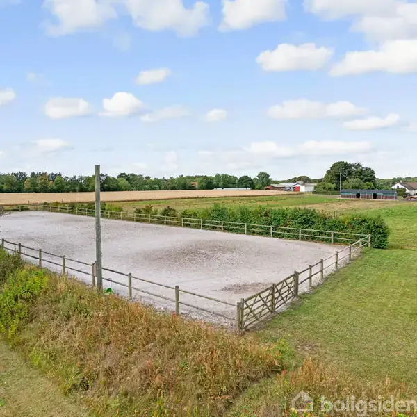 Indhegnet ridebane ligger stille omkranset af græsarealer. Baggrunden viser flade marker og et par bygninger i horisonten under en blå himmel med skyer.