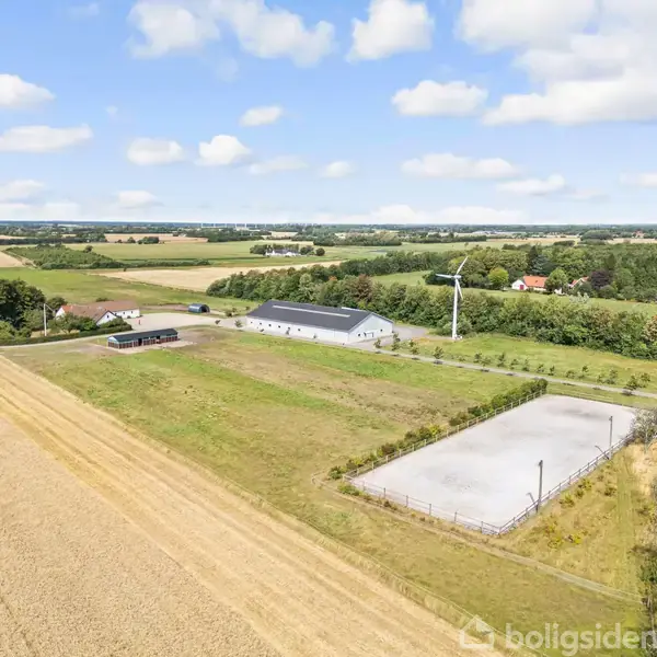En stor ladebygning står på en landlig ejendom med marker omkring sig. En vindmølle er placeret ved siden af. I baggrunden er der skov- og marklandskaber under en blå himmel.