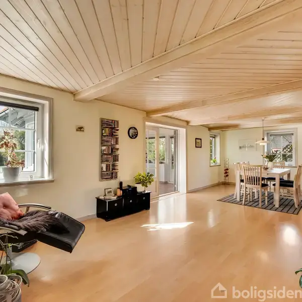 Stue med lys trægulv og loft, lænestol med pude til venstre, spisebord med stole på tæppe til højre, og vinduer med dagslys. Grønt udenfor.