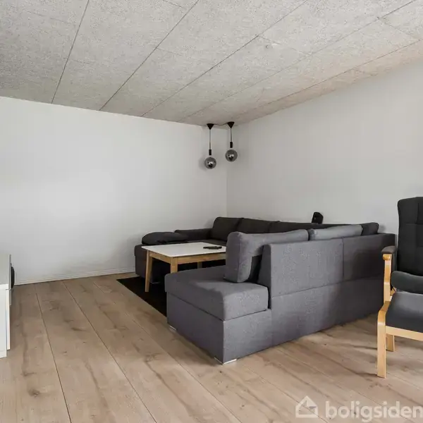 Et gråt sofaarrangement står i en stue med trælamingulv. En hvid væg har et vægmonteret TV. En lænestol står ved siden af sofaen, og et sofabord står i midten.