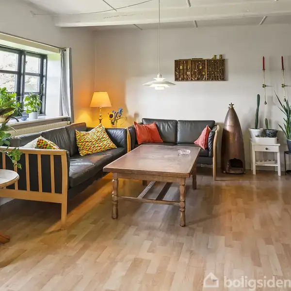 En stue med en brun sofa og farverige puder står op ad væggen, omgivet af et sofabord. Til venstre er en lampe ved et vindue. Potteplanter dekorerer rummet.