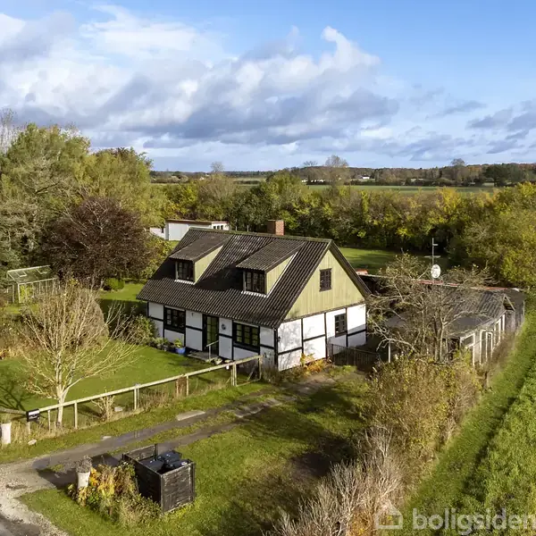 En bindingsværkshus står omgivet af frodig natur og træer, med en græsplæne foran. Det er beliggende ved en landevej på en landlig åben mark i baggrunden.
