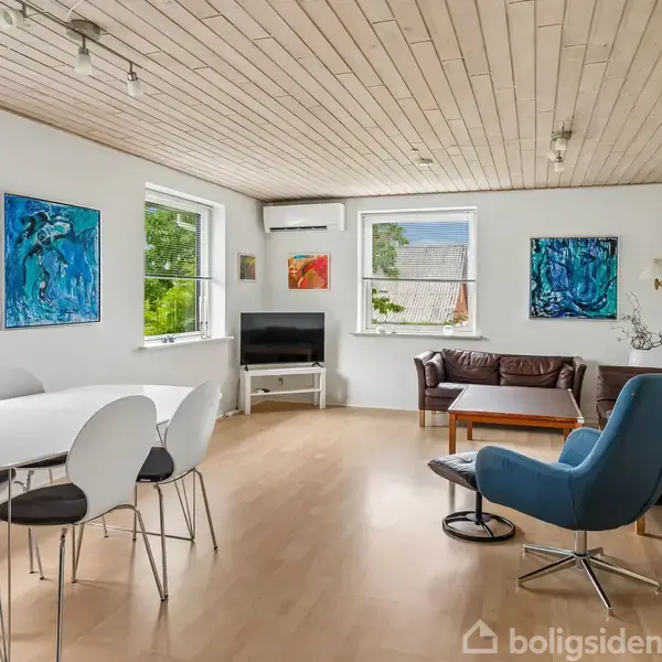 Spisestue med hvidt bord og stole. Sofa står ved væggen nær et TV. Rummet har trægulv, træloft, malerier på væggene og store vinduer med udsigt til have.