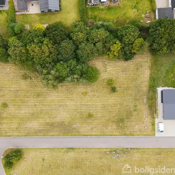 Parcel jord med græs og træer ligger ved en række huse. Til højre er et nyt hus med indkørsel. En vej løber langs bunden af billedet.