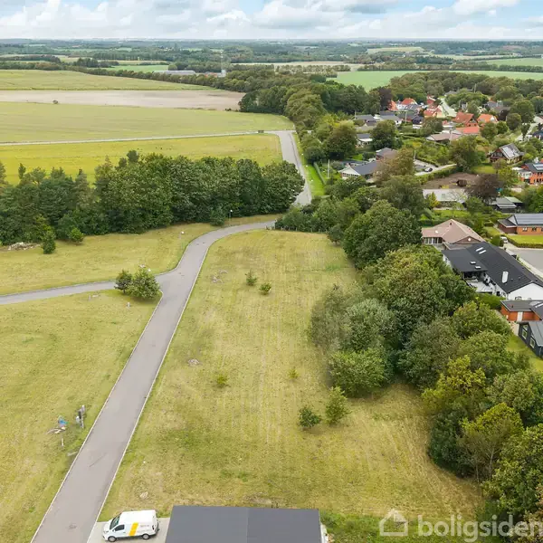 En mark med en vej snor sig gennem landskabet. Til højre er et boligområde med huse omgivet af træer. Landbrugsjord og skove ses i baggrunden under en overskyet himmel.
