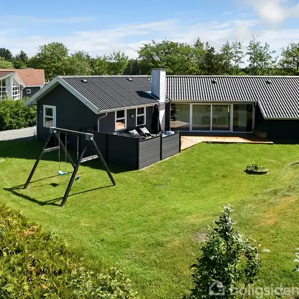 Sort, én-etagers hus med terrasse, beliggende i en velplejet have med gyngestativ. Huset har store vinduer, og området er omgivet af grønne træer og hække.
