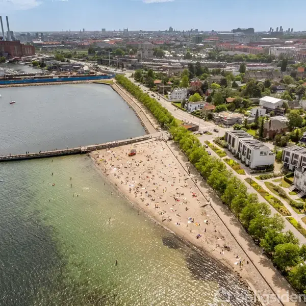Strand fyldt med mennesker nyder solen, omgivet af bygninger og træer. Havet er roligt, og i baggrunden ses en by med en industriområde og en havn.