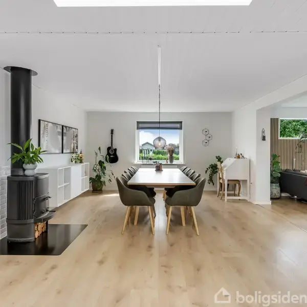 Spisebord står i et moderne, lyst rum med trægulv, omgivet af stole. En brændeovn står til venstre. Store vinduer og grønne planter skaber en indbydende atmosfære.