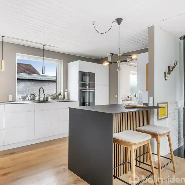 Et moderne køkken med en stor sort køkkenø; to barstole står ved øen. Til venstre ses hvide skabe og en indbygget ovn. Der hænger minimalistiske lamper og billeder på væggen. En brændeovn og planter er i baggrunden.