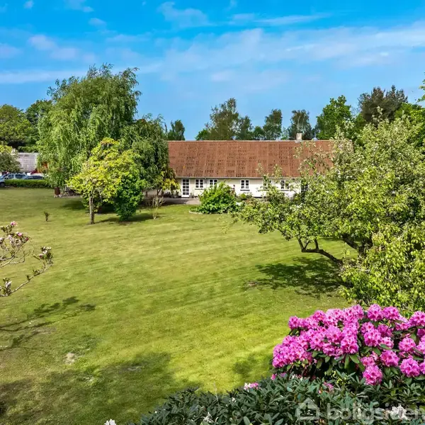 En stor have med en græsplæne strækker sig foran et traditionelt hus med rødt tag. Blomstrende buske og træer omgiver området under en klar blå himmel.