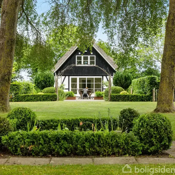 Et traditionelt, stråtækt hus med hvide vinduer står i en velplejet have. To stole er placeret på terrassen. Huset er omgivet af grønne buske og høje træer.