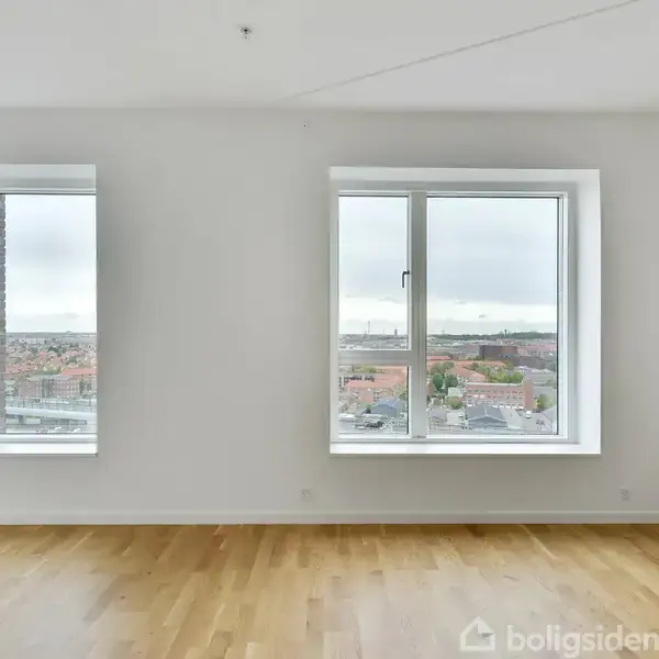 To store vinduer slipper lys ind i et lyst værelse med trægulv. Udenfor ses et bylandskab med bygninger og en delvis overskyet himmel.