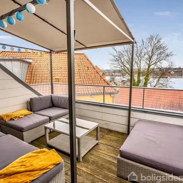 Terrasse med grå sofaer og farverig lysguirlande. Sofaerne står under en baldakin. Udsigt over hustage og vand, omgivet af træer i en bykontekst.