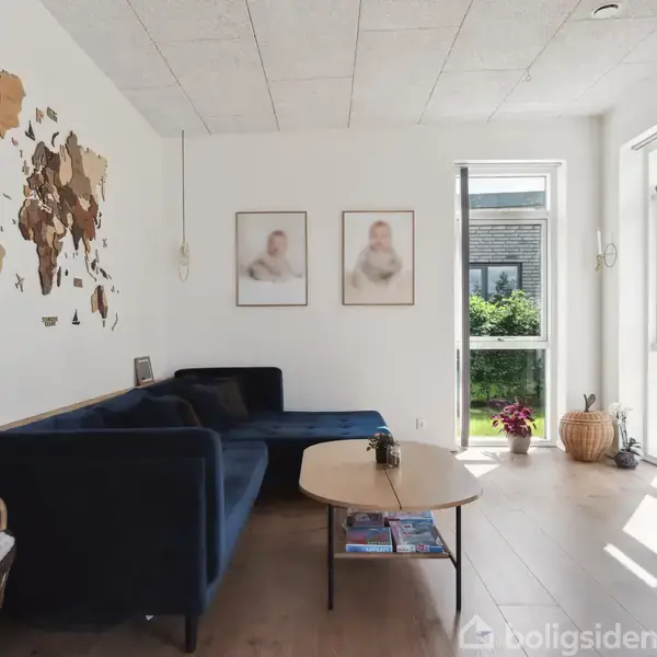 Sofa i blå farve står i stuehjørnet med et sofabord foran. På væggen hænger et verdenskort og to fotografier. Rummet er lyst med store vinduer og planter udenfor.