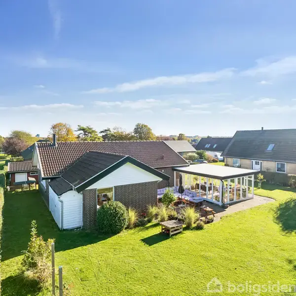En villa står stille i en velplejet have med en stor terrasse og havemøbler. Rundt omkring er der andre huse i et solrigt boligområde med blå himmel og få skyer.