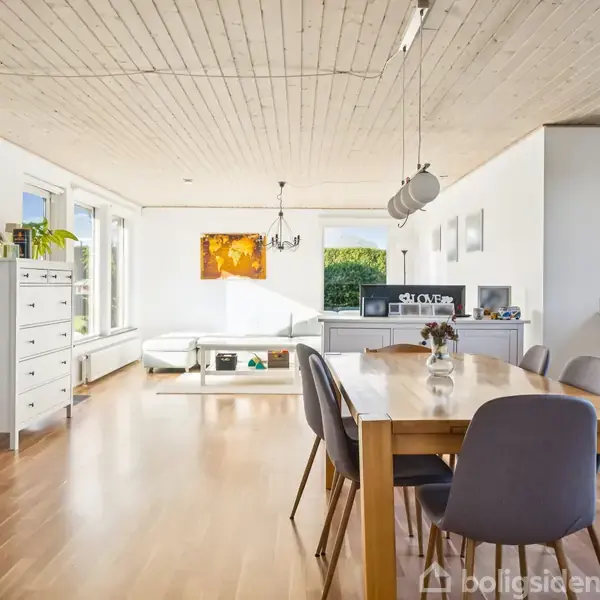 Spisebord med stole står i midten af en lys stue med parketgulv. Til venstre ses skænke og bogreoler op ad en stor glasvæg, og bagtil er en sofa ved et vindue.