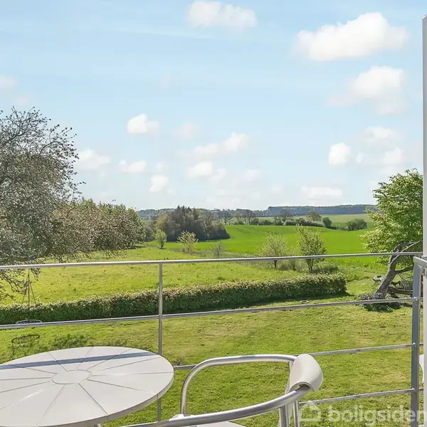 Åben dør fører ud til balkon med rundt bord og stol. I baggrunden ses en vidstrakt, grøn mark med spredte træer under en blå, skyet himmel.