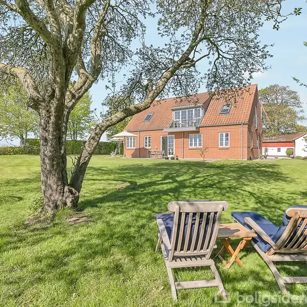 To træstole står på en grøn græsplæne foran et stort træ med en gynge. I baggrunden ses et rødt murstenshus og en klar blå himmel.
