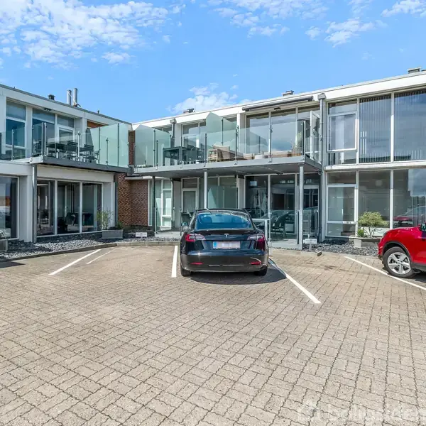 Et sort og en rød bil er parkeret foran en moderne etagebygning med store vinduer og altaner. Bygningen er omgivet af en flisebelagt parkeringsplads under en blå himmel.