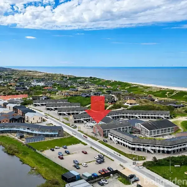 Bygning markeret med en rød pil, omringet af andre bygninger ved kystnær by. Biler parkeret på åben plads, hav i baggrunden under klar blå himmel og spredte skyer.