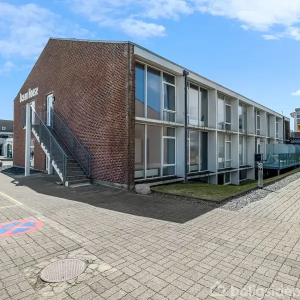 En to-etagers murstensbygning med glasfacade, trapper udenfor til venstre. Tekst på gavlen: "Ocean House". Omgivet af flisebelagt område, himmel med spredte skyer i baggrunden.