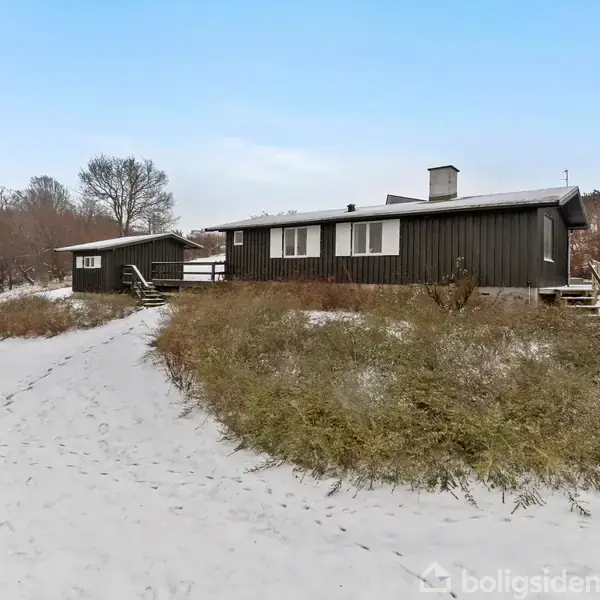 En mørk træhytte står stille på en snedækket bakke omgivet af lave buske. En sti fører op til hytten med trætrapper og uden synlige mennesker.