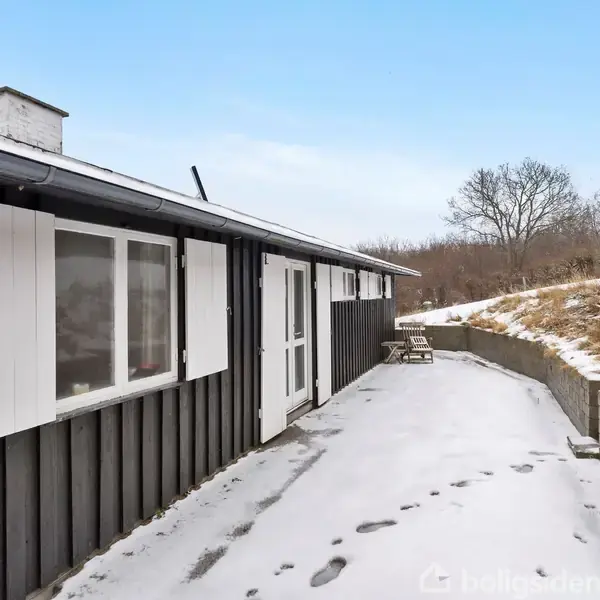 Et sort træhus med hvide skodder står langs en snedækket sti. I baggrunden er der et sneklædt græsareal og nogle træer uden blade.