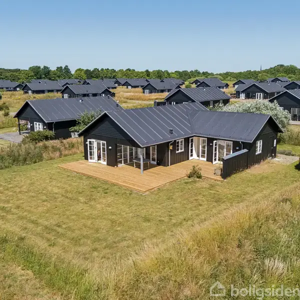 Et sort træhus står på en velholdt græsplæne, omkranset af flere lignende huse og åbne marker under en klar blå himmel.