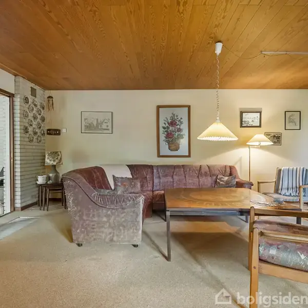Stue med hjørnesofa omkring et træbord. Væggene er dekoreret med billeder og en lampe hænger over bordet. Til venstre ses en åbning til en anden del af huset.