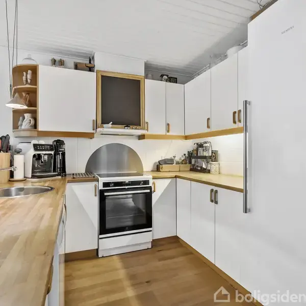 Et moderne køkken med hvide skabe, træbordplader og en indbygget ovn. En kaffemaskine og andre køkkenredskaber står på bordet. Loftslamper hænger over vasken.