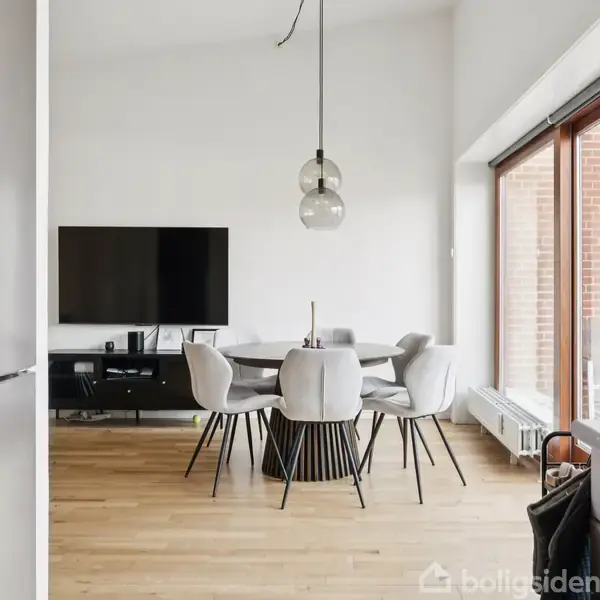 En moderne stue med trægulv har en rund spisebord med seks lyse stole. Over bordet hænger to lamper. I baggrunden er en fladskærm på en tv-bænk. Store vinduer lyser rummet.