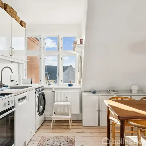 Køkken med hvide skabe og apparater, der inkluderer en ovn og vaskemaskine. Et lille step-stige står foran et vindue. Træbord og stole er ved siden af en hvid væg.