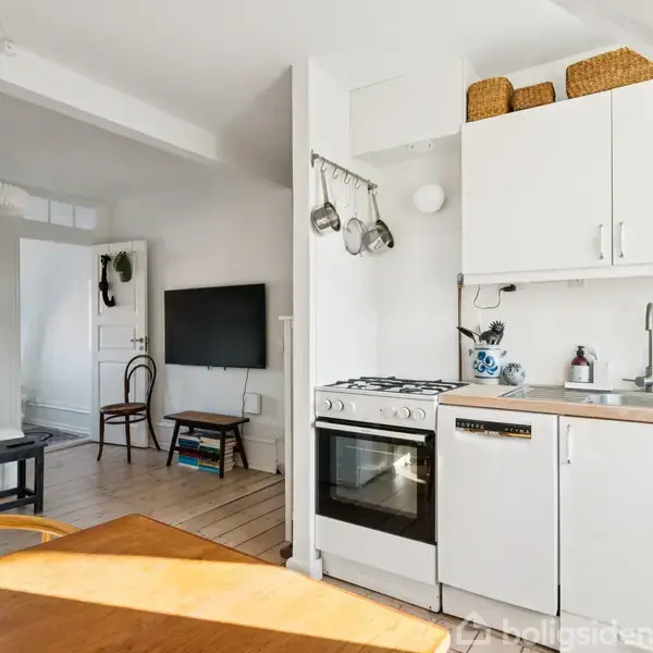 Et lyst køkken med hvidt skabarrangement og opvaskemaskine, ved siden af en ovn. Rundt bord i forgrunden. I baggrunden ses en stue med stol og vægmonteret tv.