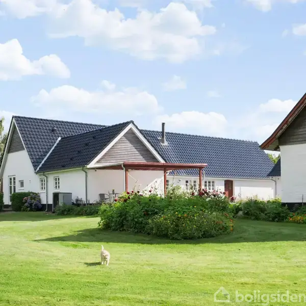 Et hvidt hus med mørkt tag står på en grøn græsplæne omgivet af blomster og buske. En kat går på plænen under en blå himmel med skyer.