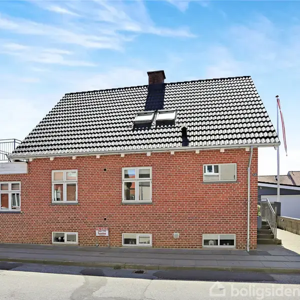 En murstensbygning står stille langs en vej. Huset har et sort-hvidt tag, og der er et hegn og en flagstang i nærheden, omgivet af andre bygninger.