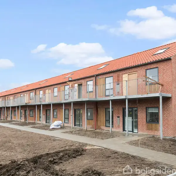 Rækkehuskompleks med to etager i rød mursten og rødt tag. Trapper og gangbroer forbinder lejlighederne. Omgivet af åben jord uden vegetation. Klar himmel i baggrunden.